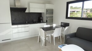 Una cocina moderna con armarios blancos, encimera negra y horno integrado. Hay una zona de comedor con mesa y sillas, y un sofá gris.