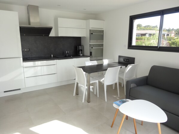 A modern kitchen with white cabinets, a black countertop, and a built-in oven. There is a dining area with a table and chairs, and a gray sofa.