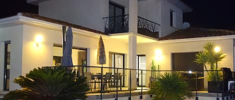 A modern two-story house with a pool, outdoor seating, and a well-lit balcony.