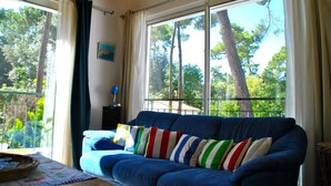 A blue sofa with striped pillows, a window with curtains, and a view of trees outside. - ROYAN Charming house in the pines 400 m from the beach, Parc district  (Saint-Georges-de-Didonne)