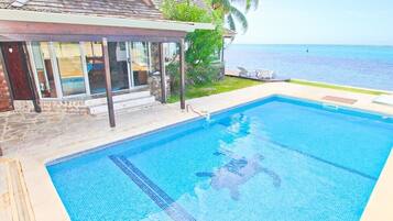 Une piscine à l’eau d’un bleu cristallin, entourée d’une terrasse en pierre et d’un espace abrité avec des portes vitrées.