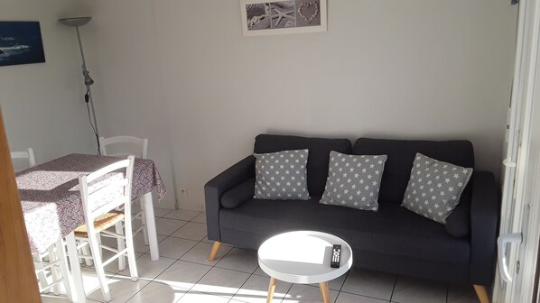 A living room with a dark sofa, three pillows, a small round table with a remote control, a white chair, and a dining table with a floral tablecloth. - Maisonnette 1Km from beaches and shops for 4 people. Pets welcome (Erdeven)