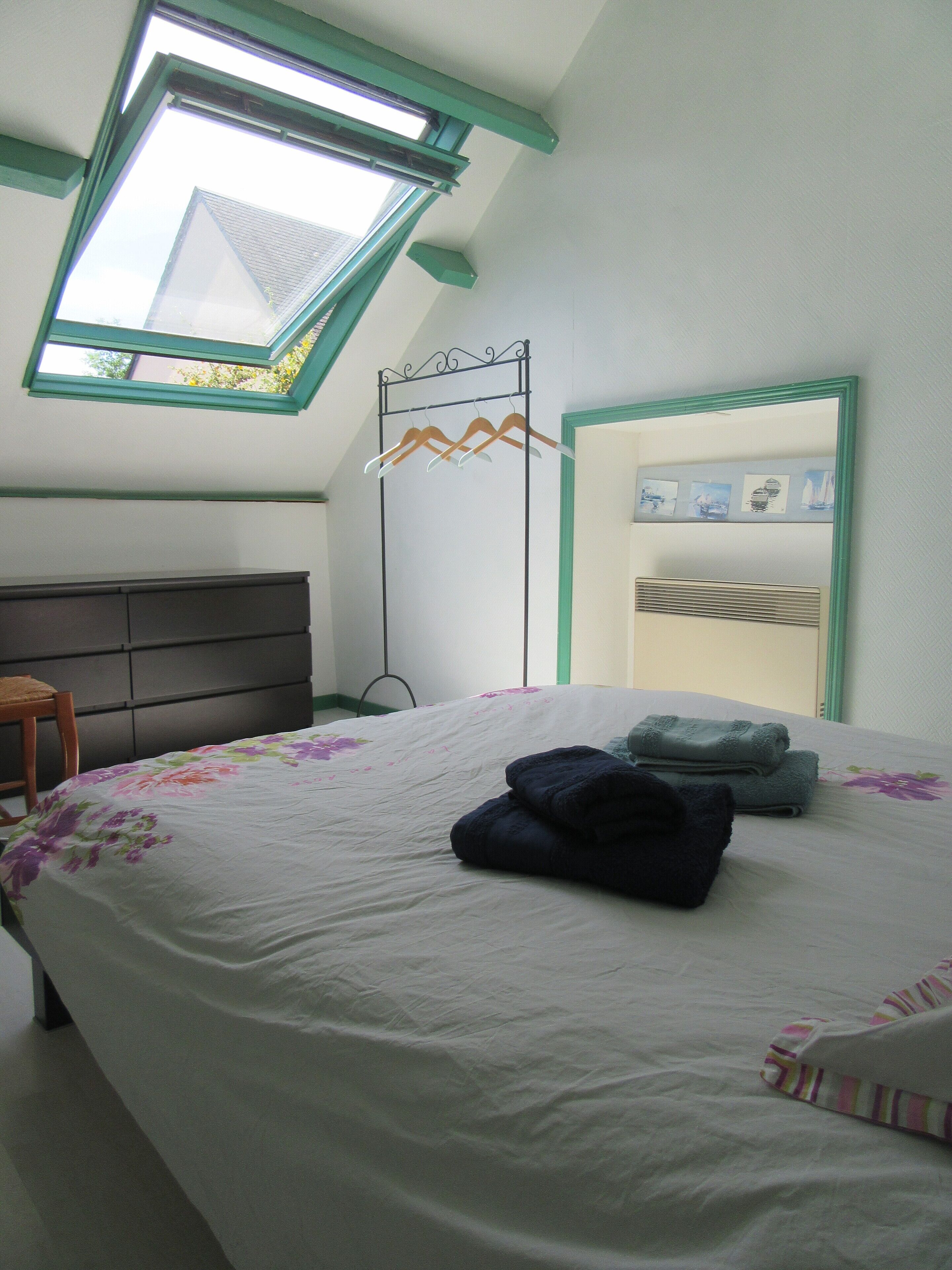 A bedroom with a skylight, a bed with a floral bedspread, a wooden chair, a bedside table, and a wall-mounted radiator.