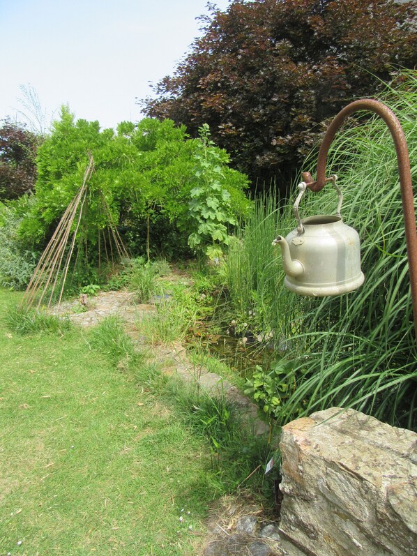 A rustic outdoor setting with a metal kettle hanging on a rusty hook, surrounded by lush greenery and a stone path. - Gîte of character near Rance near ST Malo, Dinan, Dinard (Plouer-sur-Rance)