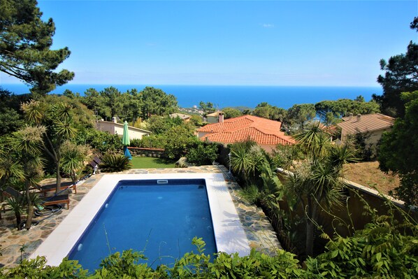 Piscina rodeada por vegetação exuberante, com vista para o mar e casas vizinhas.