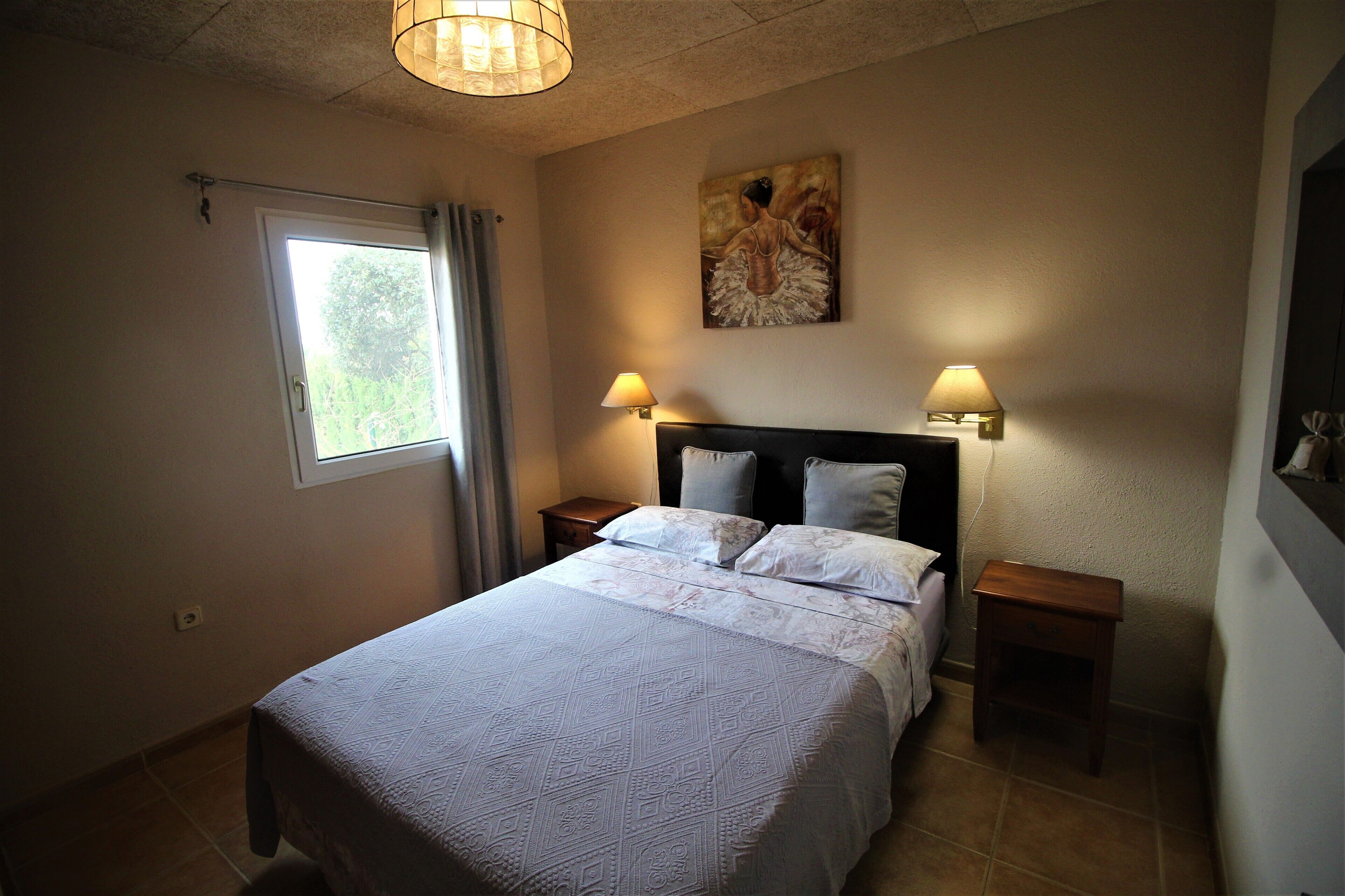 A bedroom with a large bed, two bedside tables with lamps, a window with curtains, and a painting on the wall.