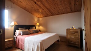 Une chambre avec un plafond en bois, un lit recouvert d’une couvre-lit blanche et rose, deux lampes de chevet, une table de chevet en bois et une commode en bois.