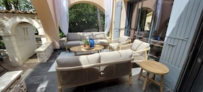 Een overdekt terras met een bank, stoelen, tafels, stenen muren en uitzicht op groen.