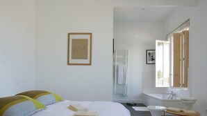 A bedroom with a large bed, a bedside table, a small stool, a window, and a bathroom with a sink and mirror.