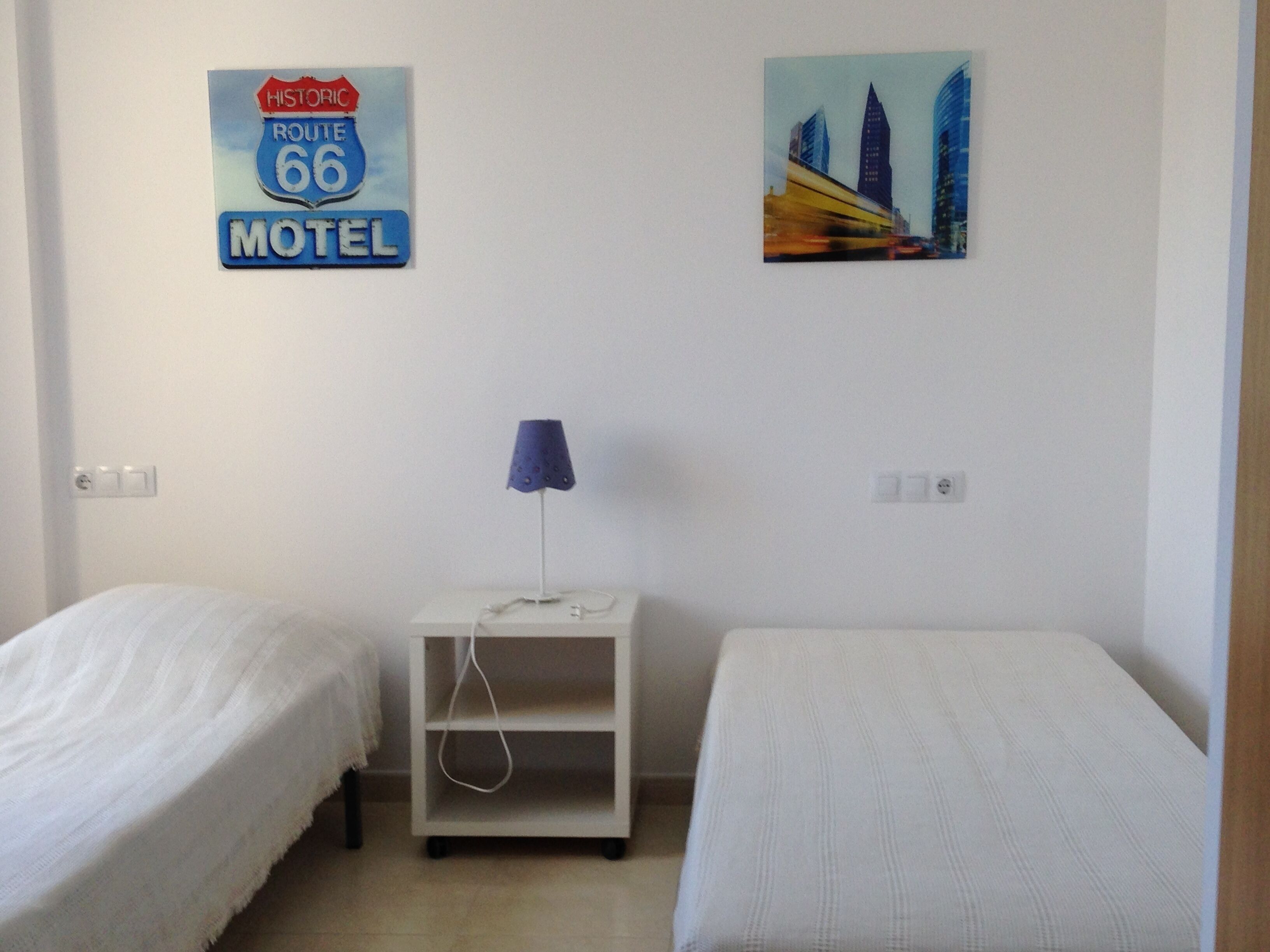 Une chambre avec deux lits simples, une table de chevet avec une lampe, un panneau mural indiquant qu’il s’agit d’un motel, et un tableau représentant un paysage urbain.