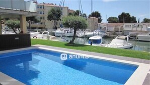 Une piscine rectangulaire à l’eau d’un bleu limpide, entourée d’une terrasse carrelée. Au fond, on aperçoit plusieurs bateaux amarrés près d’une marina, ainsi que des bâtiments dotés de balcons.
