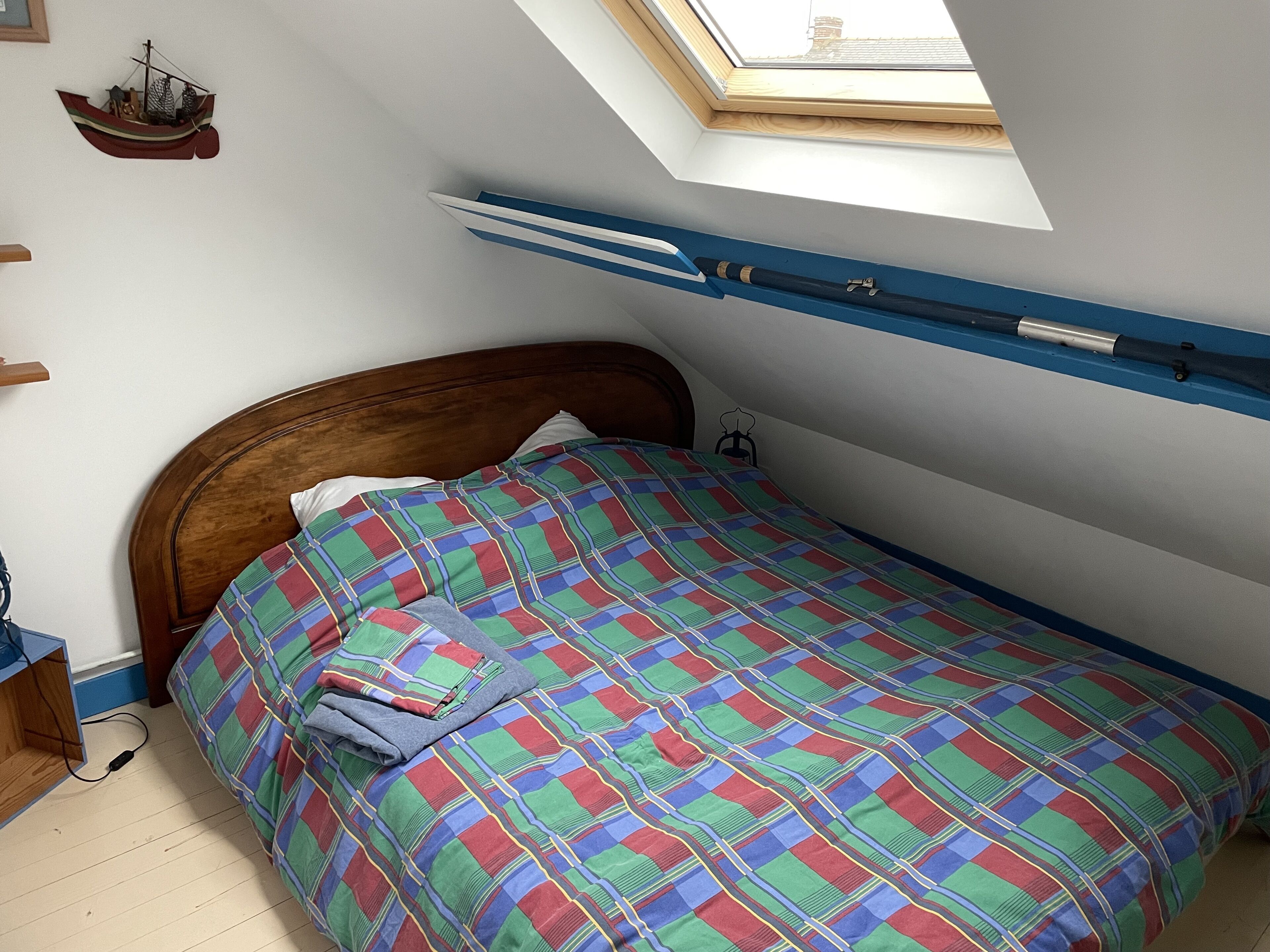 A bedroom with a sloped ceiling, a skylight, a bed with a colorful checkered bedspread, a wooden headboard, and a small ship decoration on the wall.