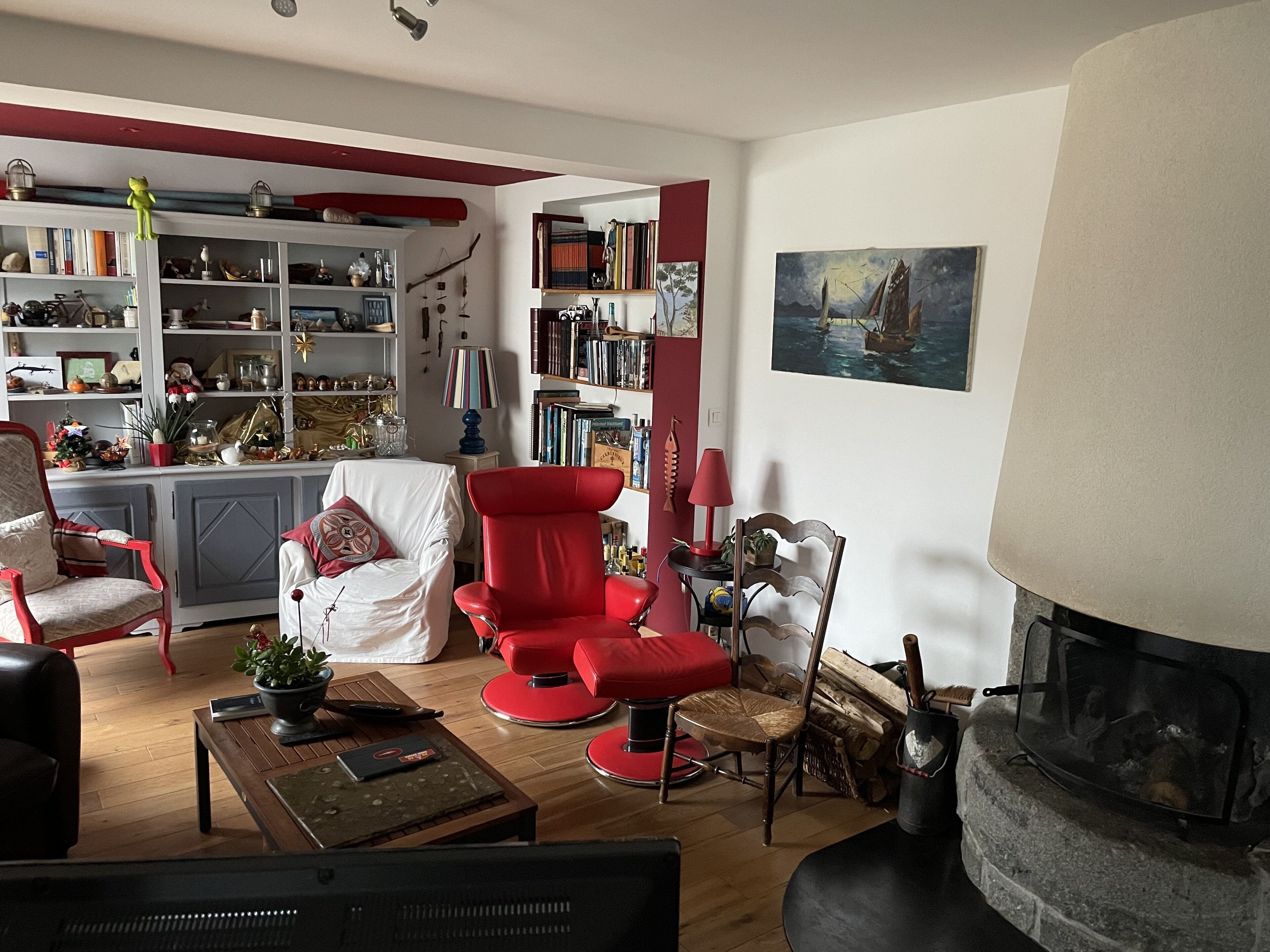 A cozy living room with a fireplace, a red armchair, a bookshelf, and a painting on the wall.