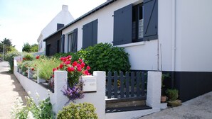 Un edificio blanco con contraventanas negras, una cerca de piquetes y diversas macetas con plantas y flores a lo largo de la fachada.