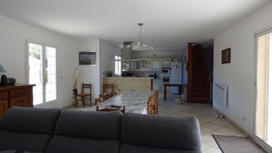 Un saló amb un sofà, taula de menjador i cadires. Al fons, es pot veure una cuina amb armaris i electrodomèstics.