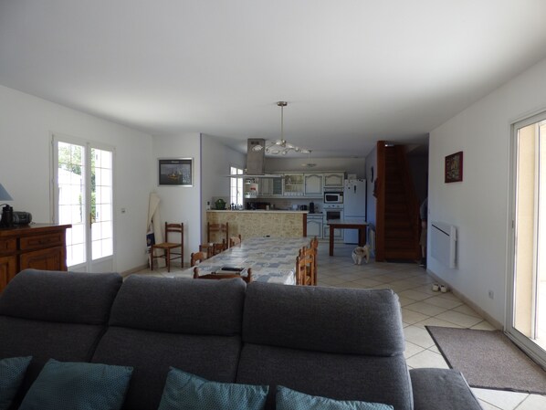 A living room with a sofa, dining table, and chairs. There is a kitchen in the background with cabinets and appliances.