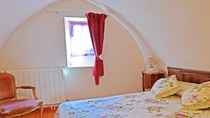 Ein Schlafzimmer mit schrägen Wänden, einem Holzstuhl, einem Bett mit floralem Bettzeug, einem Nachttisch mit Lampe und einem Fenster mit Vorhang.