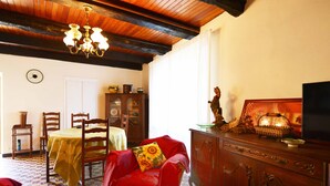 Ein Zimmer mit Holzboden, einer Kristallleuchter, einem Esstisch mit Stühlen, einem roten Sessel mit Sonnenblumenkissen, einem Holzschrank und einem Fernseher.