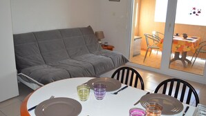 A dining area with a round table set for four, a grey sofa, and a view of an outdoor patio with a table and chairs. - Studio cabin Banyuls 4 pers, sea view, parking, WiFi (Banyuls-sur-Mer)