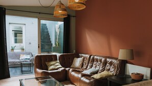 A living room with a brown leather sofa, a glass coffee table, and a lamp on a side table. There is a window with a view of a staircase and a door leading outside.