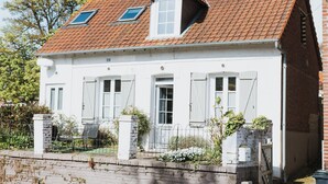 A white house with a red-tiled roof, two windows with shutters, and a small garden area with a bench and a table.
