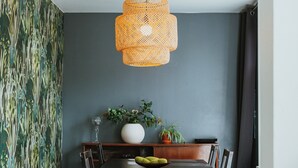 A dining area with a dark wooden table, a woven pendant light, a white vase with flowers, and a potted plant.