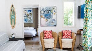 Una habitación con una cama, dos sillones de mimbre con cojines rojos y blancos, una mesa de vidrio y obras de arte en las paredes.