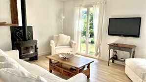Un salón con chimenea, muebles blancos, una mesa de centro de madera, un televisor y vistas a la vegetación exterior.