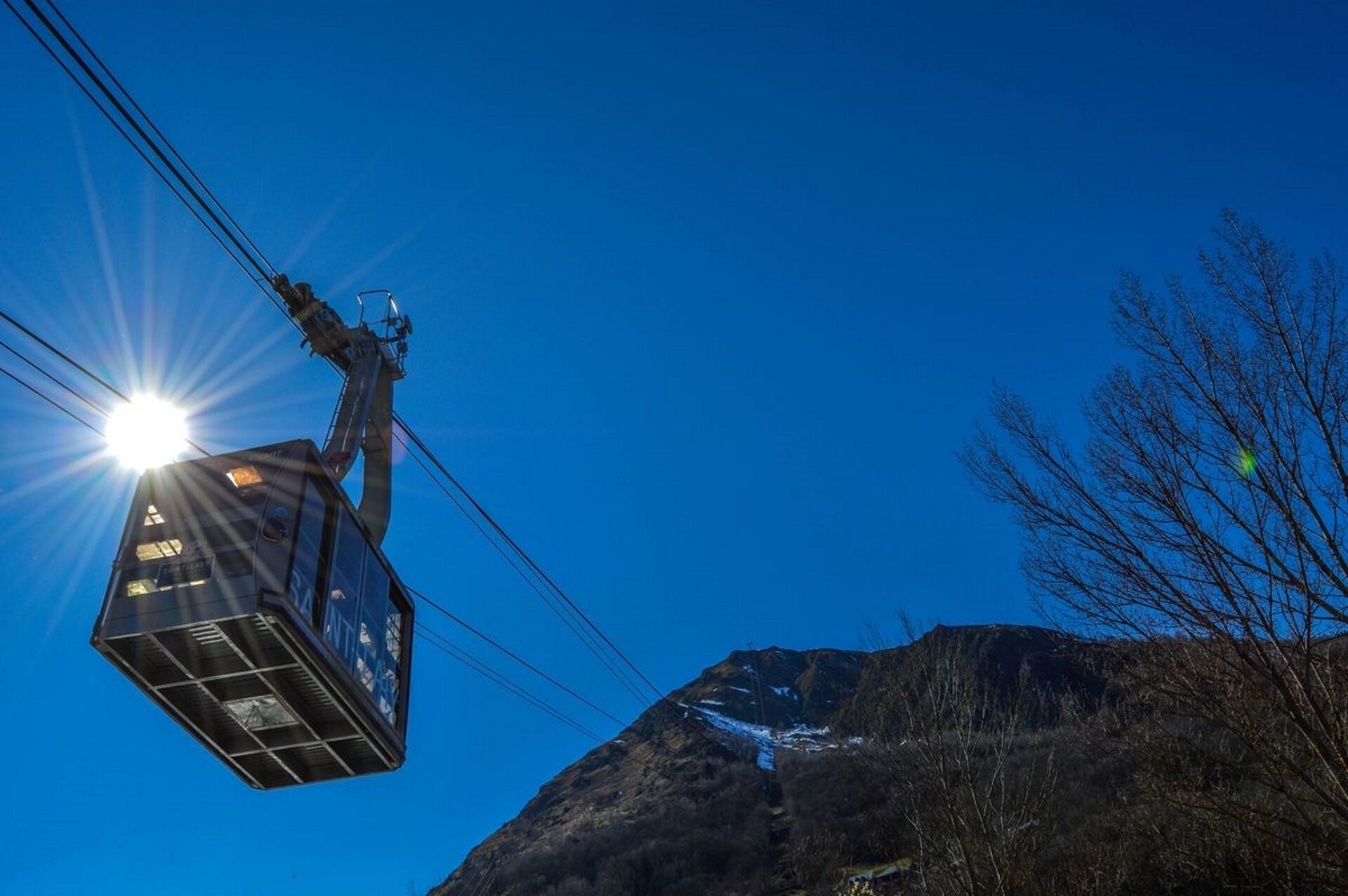 Un teleférico con un vagón iluminado por el sol, un cielo azul despejado y montañas con árboles sin hojas al fondo.