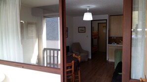 Ein Zimmer mit Holzboden, einem Esstisch mit Stühlen, einem Einzelbett, einer kleinen Küchenzeile und einem Fenster mit Vorhängen.
