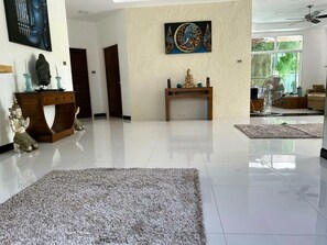 A spacious living room with a large window, a ceiling fan, and a decorative Buddha statue. - Villa Private Pool rawai (phuket rawai)