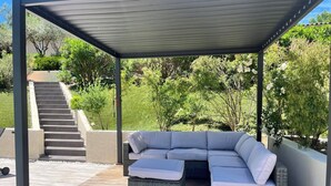 Un espacio de estar al aire libre cubierto con un sofá modular y una mesa pequeña, rodeado por una terraza de madera y una escalera que conduce a un jardín.
