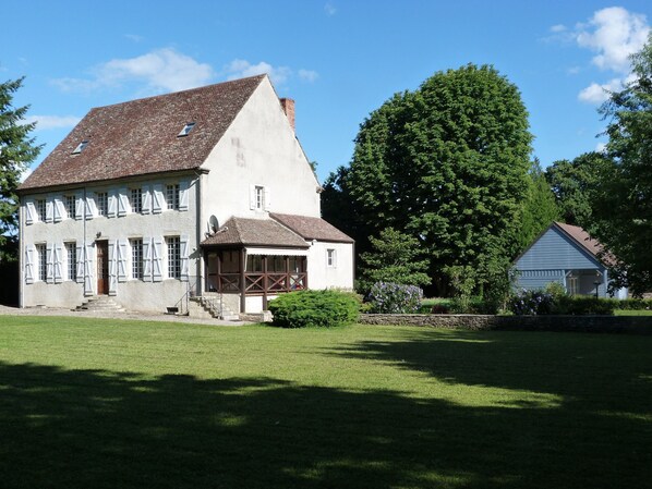 Un grand bâtiment blanc, doté d’un toit en pente, d’un petit porche et d’un grand arbre en arrière-plan.