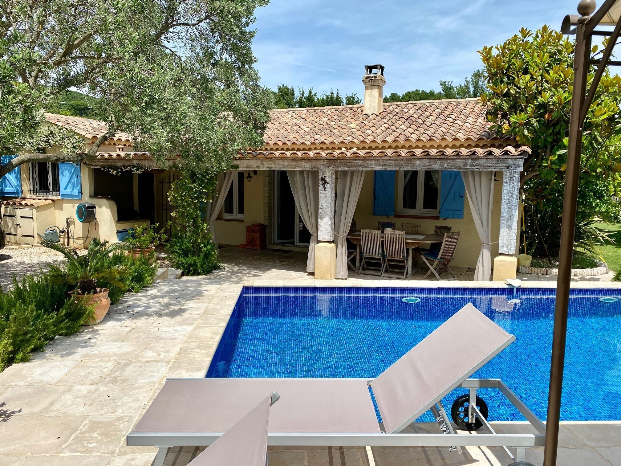 A swimming pool with a tiled deck, a covered patio area with outdoor furniture, and a house with a tiled roof and windows with blue shutters.