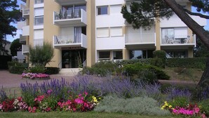 Um edifício residencial com vários andares, varandas, um jardim bem cuidado com diversas flores e um céu claro.