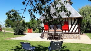 A traditional half-timbered house with a stone foundation, a red door, and a small porch with wooden furniture. - Typical cottage in Normandy Campaign (Plasnes)