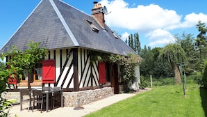 A traditional half-timbered house with a steeply pitched roof, stone foundation, and a small garden area with a table and chairs. - Typical cottage in Normandy Campaign (Plasnes)