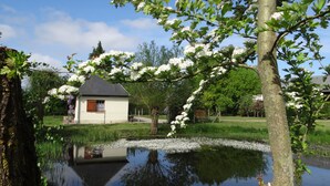 A serene lakeside scene with a small white building, a tree with white blossoms, and a clear reflection in the water. - Typical cottage in Normandy Campaign (Plasnes)