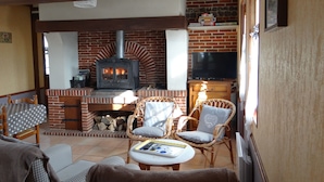 A cozy living area with a brick fireplace, wicker furniture, and a flat-screen TV. - Typical cottage in Normandy Campaign (Plasnes)