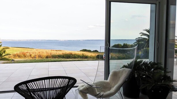 A modern room with a large glass door leading to a terrace with a view of the sea and grassy fields. - Vue Mer - Contemporary artist's house ★★★★ - Crozon - GR34 - Bretagne (Telgruc-Sur-Mer)