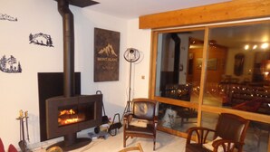 A cozy living room with a fireplace, wooden furniture, and a large window offering a view of the interior. - House for rent in the Haut Jura (Longchaumois)