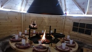 A wooden interior with a central fire, round tables with plates and cups, and a window on the right wall. - House for rent in the Haut Jura (Longchaumois)