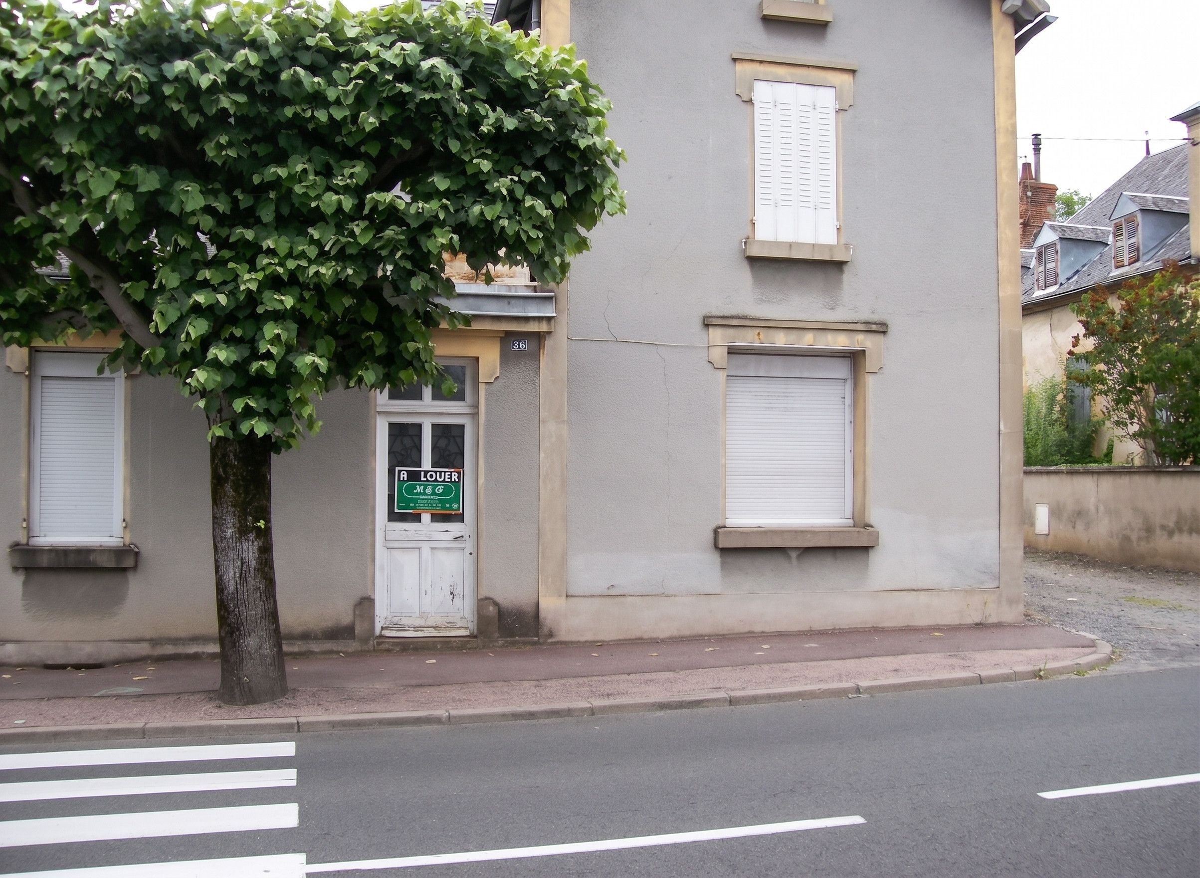 Un edificio con puerta y contraventanas blancas, un letrero verde y un árbol en primer plano.