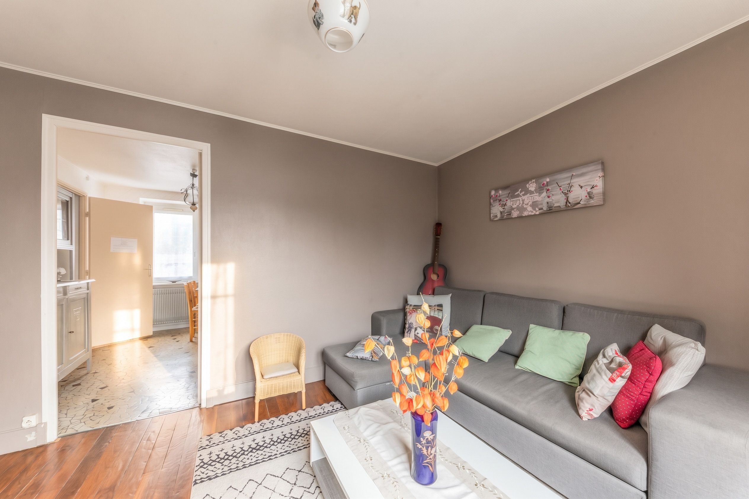 Een woonkamer met een grijze bank, een rieten stoel, een vaas met oranje bloemen en een schilderij aan de muur.