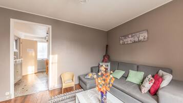 Een woonkamer met een grijze bank, een rieten stoel, een vaas met oranje bloemen en een schilderij aan de muur.