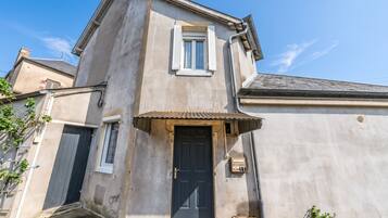 Un bâtiment à deux étages avec une porte d’entrée sombre, une petite fenêtre avec des volets et une boîte électrique visible sur le mur.