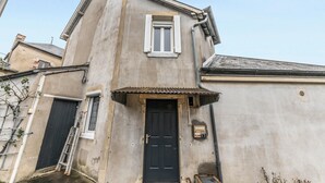 Un edificio di due piani con una porta nera, una piccola finestra con persiane e una scala di metallo appoggiata al muro.