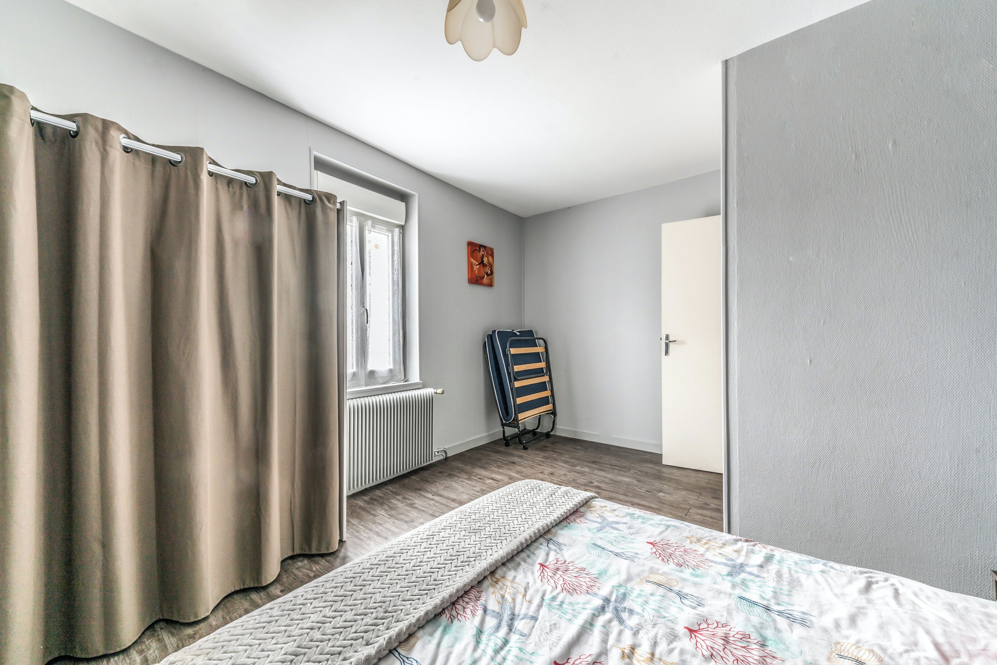 Ein Schlafzimmer mit Bett, Vorhängen, Heizkörper, Klappstuhl und Tür.