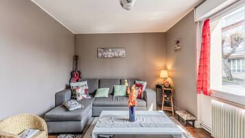 A living room with a grey sofa, a coffee table, a wicker chair, a guitar, and a window with red curtains.