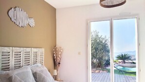 Une chambre avec un grand lit, une tête de lit en bois, un vase contenant des fleurs séchées et une vue sur une piscine et la verdure, visible à travers une porte-fenêtre coulissante.